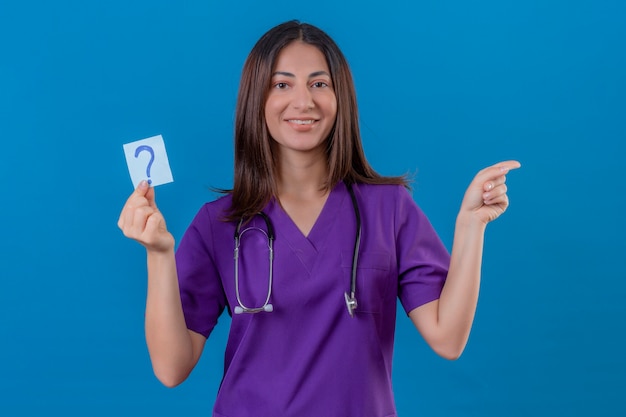 infermiera donna in uniforme medica e con uno stetoscopio tenendo la carta promemoria con un punto interrogativo molto felice indicando con la mano e il dito a lato in piedi sul blu