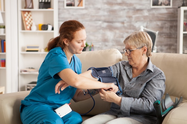 Infermiera femminile che utilizza un dispositivo digitale per la pressione sanguigna su una donna anziana in casa di cura.