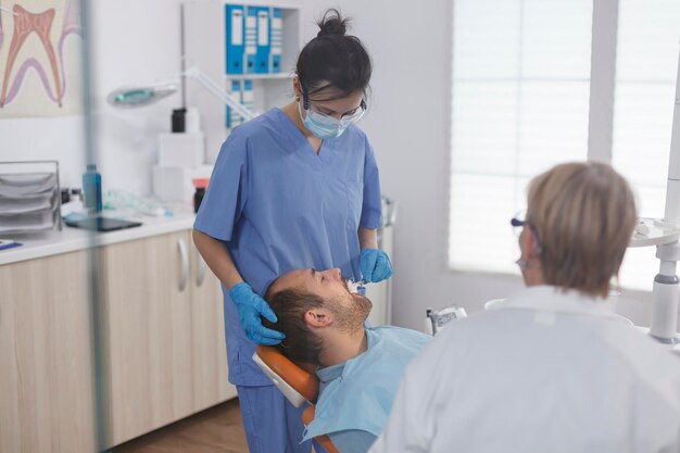 Infermiera odontoiatrica che applica l'anestesia con un batuffolo di cotone prima della procedura stomatologica in studio ortodontico. Equipe medica che discute il trattamento contro l'infezione della carie durante l'esame di odontoiatria