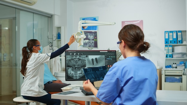 Infermiera stomatologa che confronta le radiografie guardando il computer, mentre il medico specialista con maschera facciale parla all'uomo con mal di denti seduto sulla sedia stomatologica preparando gli strumenti per la chirurgia