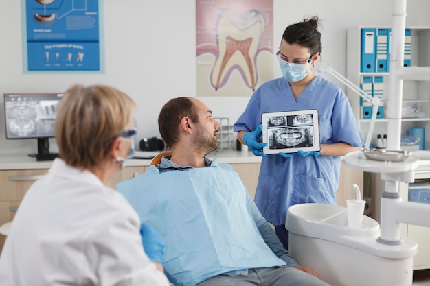 Infermiere stomatologo con maschera facciale che tiene computer tabet che spiega la radiografia dei denti al paziente malato durante la consultazione di odontoiatria nella stanza dell'ufficio stomatologico. Team che lavora al trattamento della carie