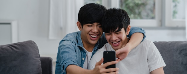 Influencer asiatico Coppia gay vlog a casa. Gli uomini asiatici LGBTQ si rilassano felici utilizzando la tecnologia di telefonia mobile record di stile di vita video vlog upload nei social media mentre giace divano nel soggiorno.