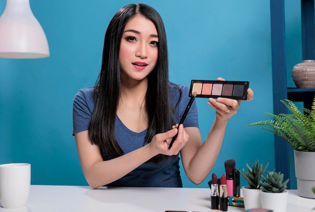 Influencer dei social media che tiene la tavolozza del trucco che mostra alla fotocamera mentre fa un tutorial di bellezza che pubblicizza un prodotto cosmetico. Creatore di contenuti asiatico che registra una recensione cosmetica per il canale di vlogging