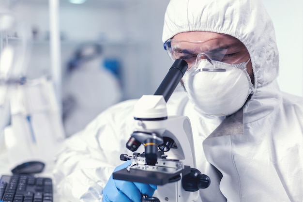 Ingegnere chimico con gli occhiali che conduce indagini sanitarie al microscopio. Scienziato in tuta protettiva seduto sul posto di lavoro utilizzando la moderna tecnologia medica durante l'epidemia globale.