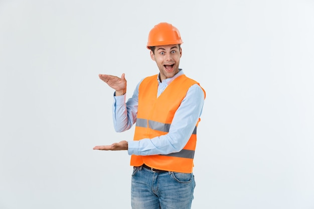 Ingegnere della barba felice che tiene la mano sul lato e spiega qualcosa, ragazzo che indossa maglietta e jeans caro con gilet giallo e casco arancione, isolato su sfondo bianco.