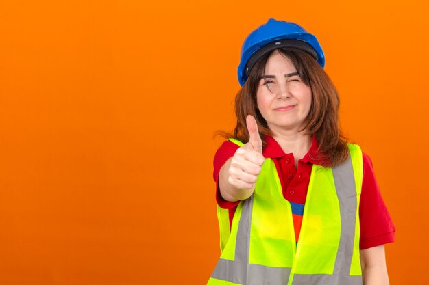 Ingegnere della donna in maglia della costruzione e casco di sicurezza che sbattono le palpebre mostrando pollice su che sorride sopra la parete arancio isolata