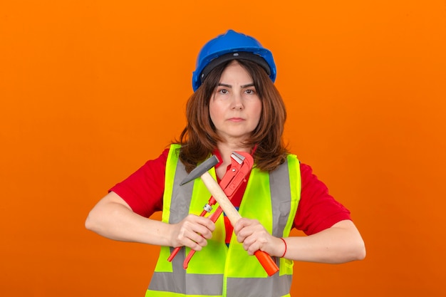 Ingegnere donna che indossa giubbotto di costruzione e casco di sicurezza con martello e chiave regolabile nelle mani simbolo di uguaglianza con uomini in piedi su muro arancione isolato