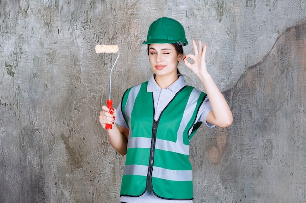 Ingegnere donna con casco verde che tiene in mano un rullo di rifinitura per la pittura murale e mostra un segno positivo con la mano