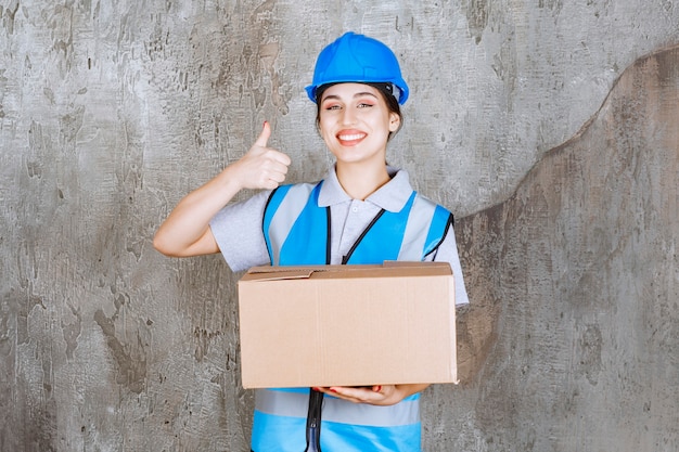 Ingegnere donna in uniforme blu e casco che tiene un pacco di cartone e mostra un segno positivo con la mano