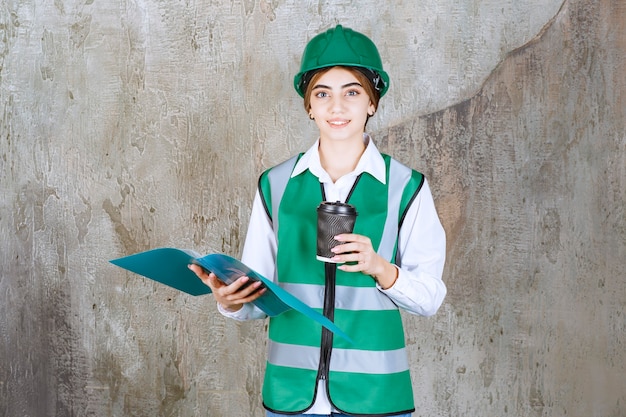 Ingegnere donna in uniforme verde e casco in possesso di una cartella di progetto verde e una tazza di bevanda