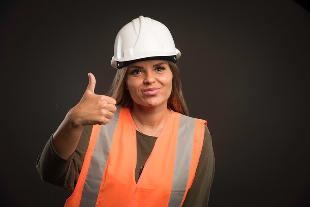 Ingegnere femminile che indossa un casco e un ingranaggio bianchi e che dà pollice sul segno.