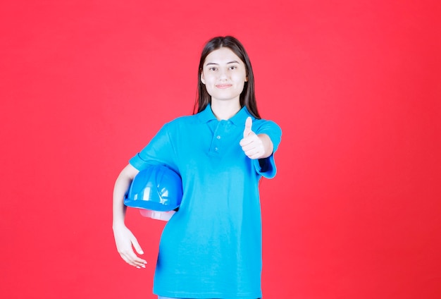 Ingegnere femminile che tiene il casco blu e che mostra il segno positivo della mano