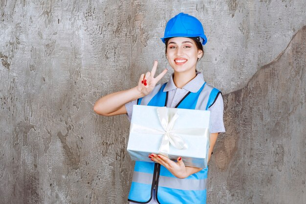 Ingegnere femminile in uniforme blu e casco che tiene una confezione regalo blu e che mostra il segno positivo della mano.