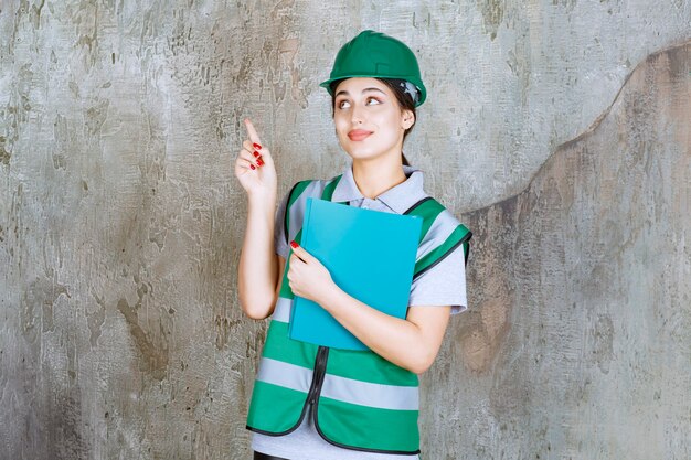 Ingegnere femminile in uniforme verde e casco che tiene una cartella di progetto blu e indica l'edificio.