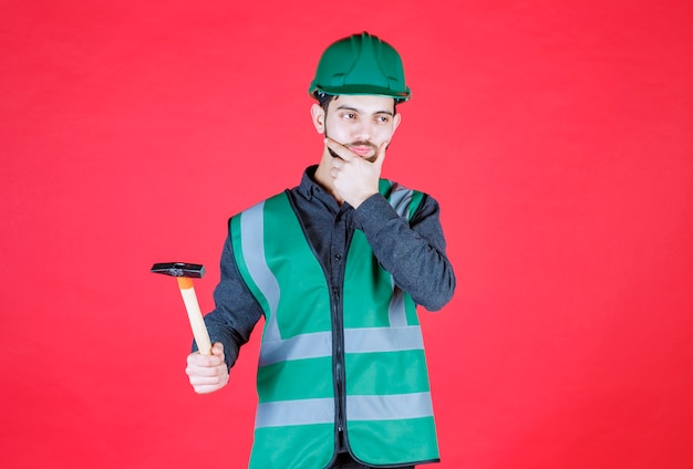 Ingegnere in uniforme verde e casco che tiene un'ascia di legno e pensa.