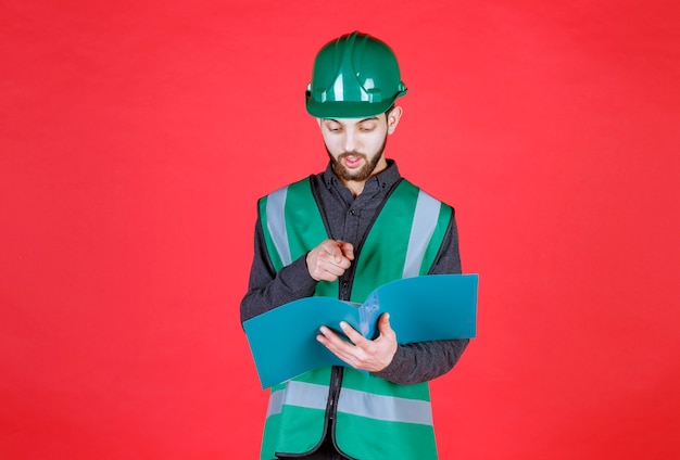 Ingegnere in uniforme verde e casco in possesso di una cartella blu, leggendo e apportando correzioni.