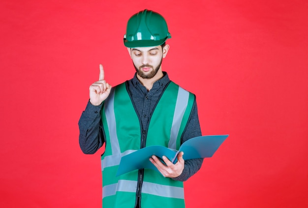 Ingegnere in uniforme verde e casco in possesso di una cartella blu, leggendo e apportando correzioni.