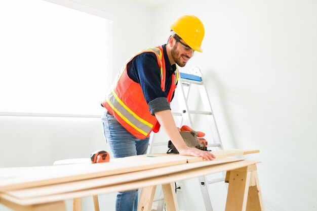 Ingegnere maschio felice con un casco di sicurezza che sorride mentre taglia un pannello di legno con una macchina elettrica