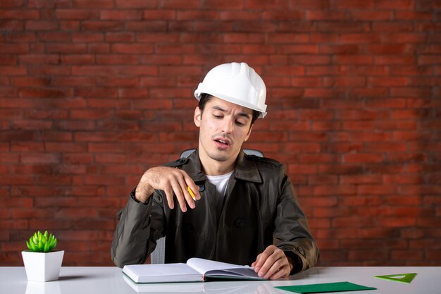 Ingegnere maschio vista frontale seduto dietro il suo posto di lavoro in casco bianco documento affari proprietà corporative lavoro agenda costruttore appaltatore piano