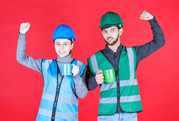 Ingegneri maschi e femmine con il casco che tiene tazze blu e verdi e che mostra il segno di soddisfazione.