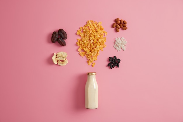 Ingredienti per la colazione muesli fatta in casa. Bottiglia di latte fresco, cereali e frutta secca come mela, cocco, uva intorno isolato su sfondo rosa. Preparare il muesli. Perfetto spuntino nutriente