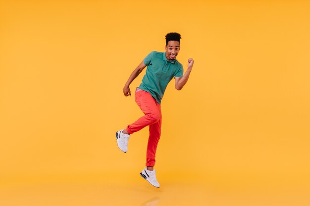 Inquadratura a figura intera di un ragazzo nero ispirato che balla in pantaloni rossi. Foto dell'interno dell'uomo africano spensierato che gode.
