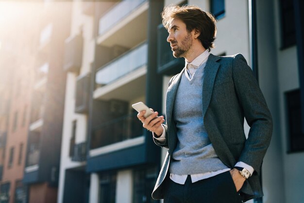 Inquadratura dal basso dell'uomo d'affari che utilizza il telefono cellulare mentre è in piedi sulla strada e distoglie lo sguardo.