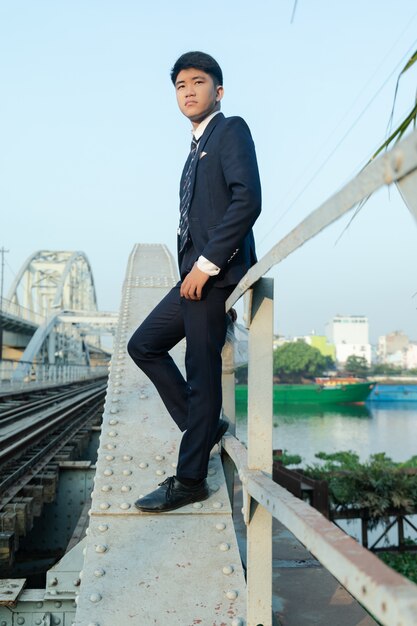 Inquadratura dal basso di un giovane uomo asiatico in un vestito che si appoggia sulle ringhiere del ponte