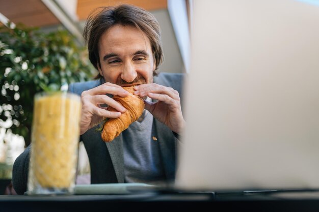 Inquadratura dal basso di un giovane uomo d'affari che mangia croissant mentre è seduto nella caffetteria all'aperto