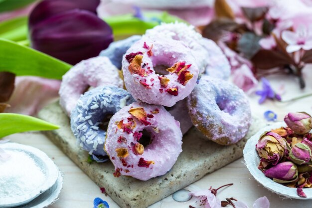 Inquadratura dall'alto di alcune ciambelle vegane blu e viola circondate da fiori su un tavolo