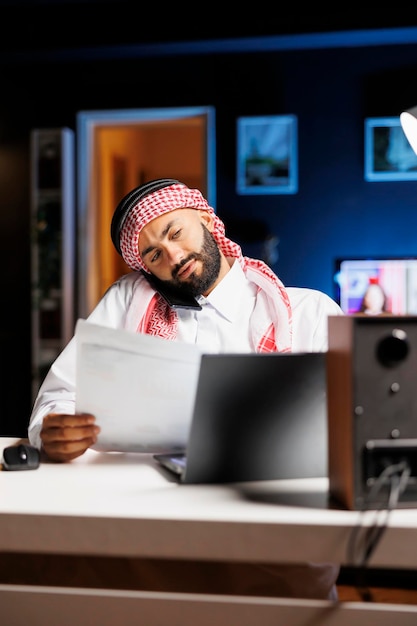 Inquadratura dettagliata di un uomo arabo che utilizza un laptop e un dispositivo mobile per la comunicazione e la ricerca, dimostrando competenza nella tecnologia. Ragazzo musulmano che fa multitasking, parla al cellulare e confronta appunti.