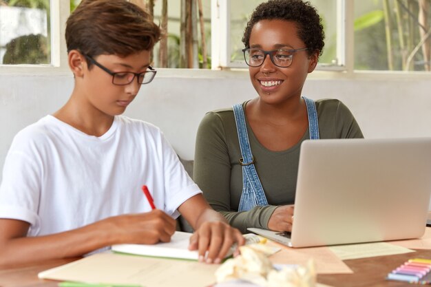 Inquadratura di studenti adolescenti multietnici coinvolti nel processo di lavoro, sfogliare le informazioni sul computer portatile portatile, scrivere idee per il lavoro di progetto nel blocco note, sedersi al desktop, controllare il messaggio di reddito