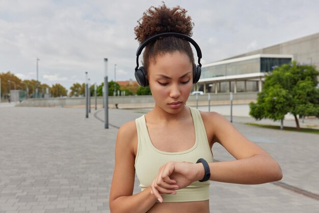 Inquadratura in vita di donna sportiva con capelli ricci pettinati utilizza smartwatch controlla le calorie bruciate ascolta musica tramite le cuffie indossa il top corto pone all'aperto allo stadio impegnato in attività sportive attive