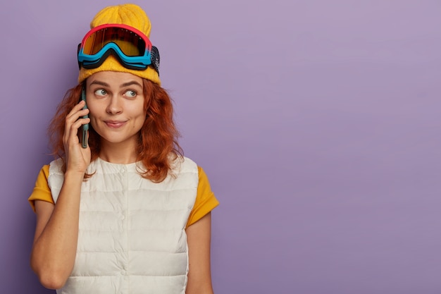 Inquadratura orizzontale della bella giovane donna allo zenzero sulla stazione sciistica, fa una telefonata, indossa un cappello giallo e un giubbotto bianco, si trova al coperto sopra la parete viola