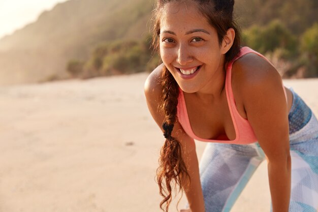 Inquadratura orizzontale della donna allegra fitness ha la pelle lentigginosa, treccia scura, vestita con abiti sportivi, sorriso piacevole