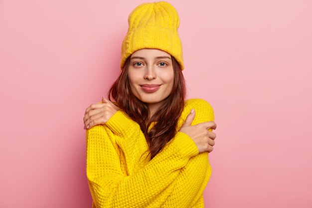 Inquadratura orizzontale della giovane donna attraente si abbraccia, ha i capelli lunghi scuri, sguardo tenero, indossa un maglione e un cappello invernali gialli, pone su sfondo rosa studio.