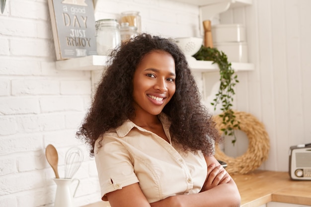 Inquadratura orizzontale di bella giovane donna mulatta nera in camicia beige che attraversa le braccia sul petto, guardando con un sorriso a trentadue denti fiducioso, in posa all'interno contro l'interno della cucina elegante