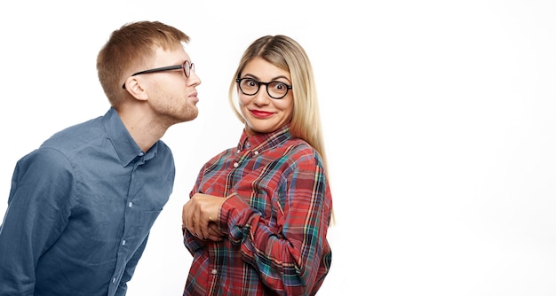 Inquadratura orizzontale di divertente ragazzo nerd con barba e occhiali chiudendo gli occhi e le labbra imbronciate come se stesse per baciare attraente giovane donna che si sente persa, guardando con un sorriso confuso