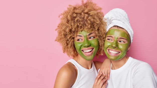 Inquadratura orizzontale di donne felici che applicano maschere nutrienti verdi sul viso guardano con gioia lontano stando vicine l'una all'altra isolate su sfondo rosa spazio vuoto per la copia del tuo contenuto pubblicitario