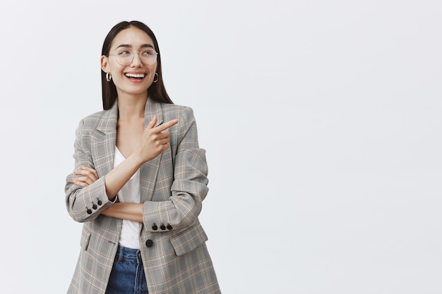 Inquadratura orizzontale di elegante donna femminile in occhiali e orecchini alla moda, ridendo di piacere e felicità mentre guarda e indica nell'angolo in alto a destra, divertendosi sul muro grigio