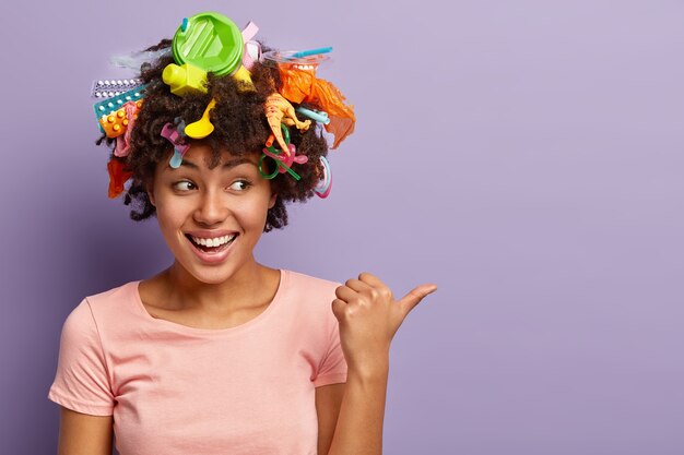 Inquadratura orizzontale di felice donna dalla pelle scura con lettiera nei capelli, punta il pollice da parte, dimostra lo spazio della copia, ride positivamente, essendo volontaria attiva, indossa una maglietta casual. Immondizia e riciclaggio