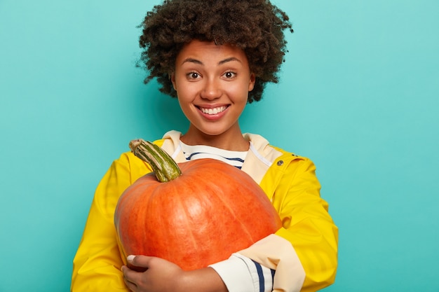 Inquadratura orizzontale di felice donna dalla pelle scura tiene maturi vegetali autunnali, abbraccia la grande zucca, ha un sorriso a trentadue denti, indossa un impermeabile giallo