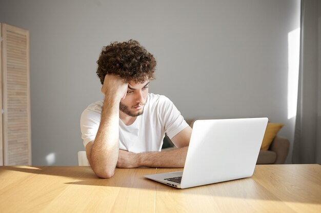 Inquadratura orizzontale di giovane freelance maschio con la barba lunga frustrato in maglietta bianca utilizzando un dispositivo elettronico per lavoro a distanza, avendo un aspetto stanco o annoiato mentre si lavora su un progetto urgente la mattina presto