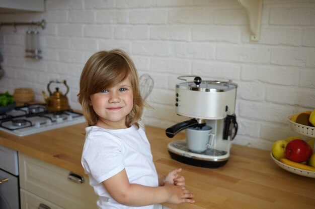 Inquadratura orizzontale di ragazzino carino spensierato di età prescolare che indossa una maglietta bianca con sguardo felice, in piedi al tavolo della cucina, facendo il caffè per suo padre. Infanzia spensierata e concetto di cucina