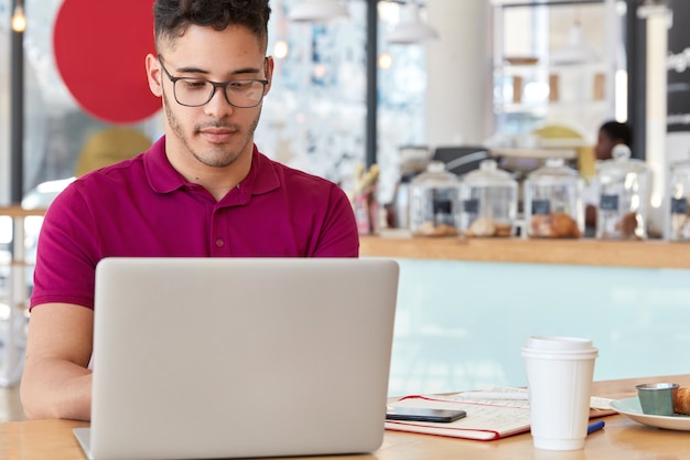 Inquadratura orizzontale di un uomo attraente con gli occhiali, guarda il video sul suo laptop, scrive sulla tastiera, i modelli in caffetteria, beve il caffè, si prepara per una conversazione di lavoro Concetto di rete