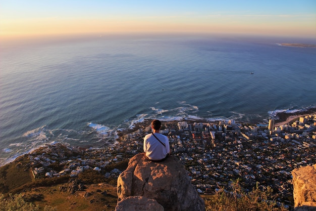 Inquadratura orizzontale di un uomo seduto sul bordo della roccia e guardando la città costiera