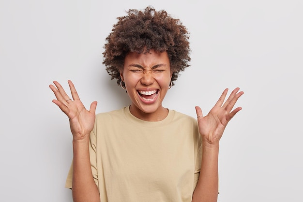 Inquadratura orizzontale di una donna riccia felice stupita che tiene i palmi delle mani sollevati reagisce a notizie fantastiche ridacchia vestita positivamente con una maglietta marrone casual isolata su sfondo bianco Concetto di reazioni umane