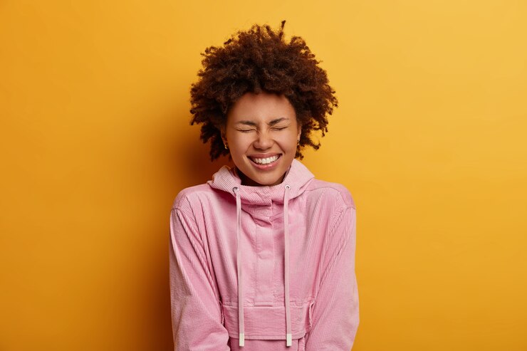 Foto gratuito inquadratura orizzontale di una giovane donna dalla pelle scura che ride di qualcosa di divertente, ridacchia per uno scherzo esilarante, si rallegra di eventi positivi, indossa una felpa con cappuccio roseo, modelli al coperto. persone, felicità, concetto di stile di vita