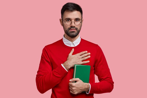 Inquadratura orizzontale di uno studente universitario maschile serio con la barba lunga promette di studiare sodo, tiene un libro di testo verde, indossa un maglione rosso elegante