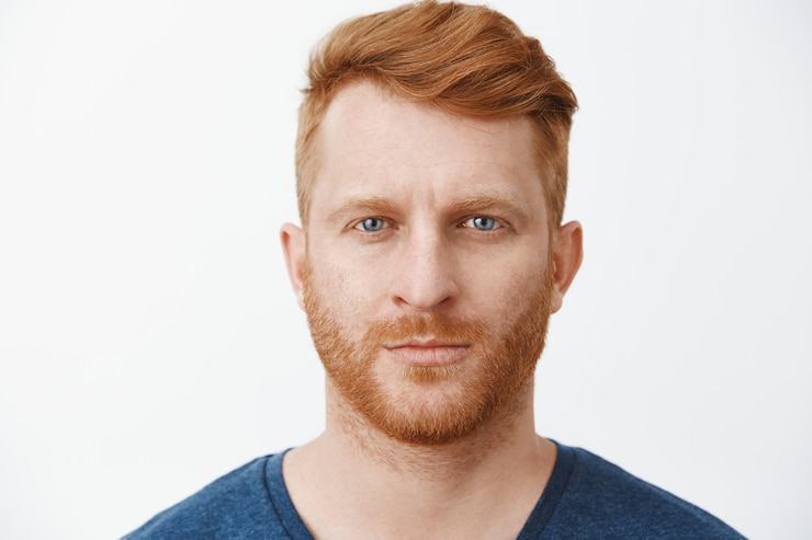 Foto gratuito inquadratura ravvicinata di un bell'uomo europeo adulto dall'aspetto serio con i capelli rossi e la barba che fissa con espressione concentrata e determinata, in piedi in posa rigorosa sul muro grigio